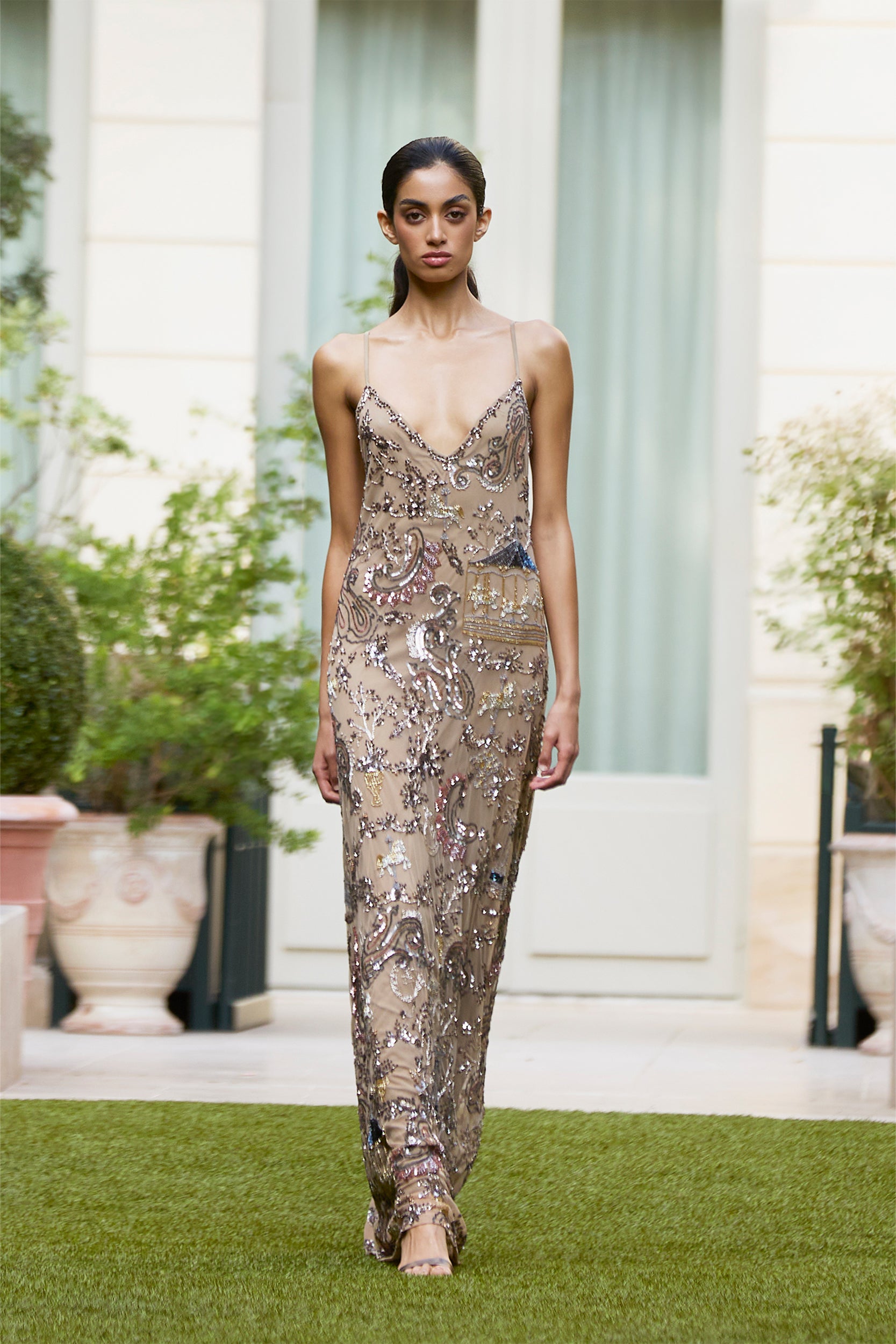 A woman models a sleeveless, shimmering, floor-length gown with intricate sequin details, walking on grass amid lush greenery and a building in the background.