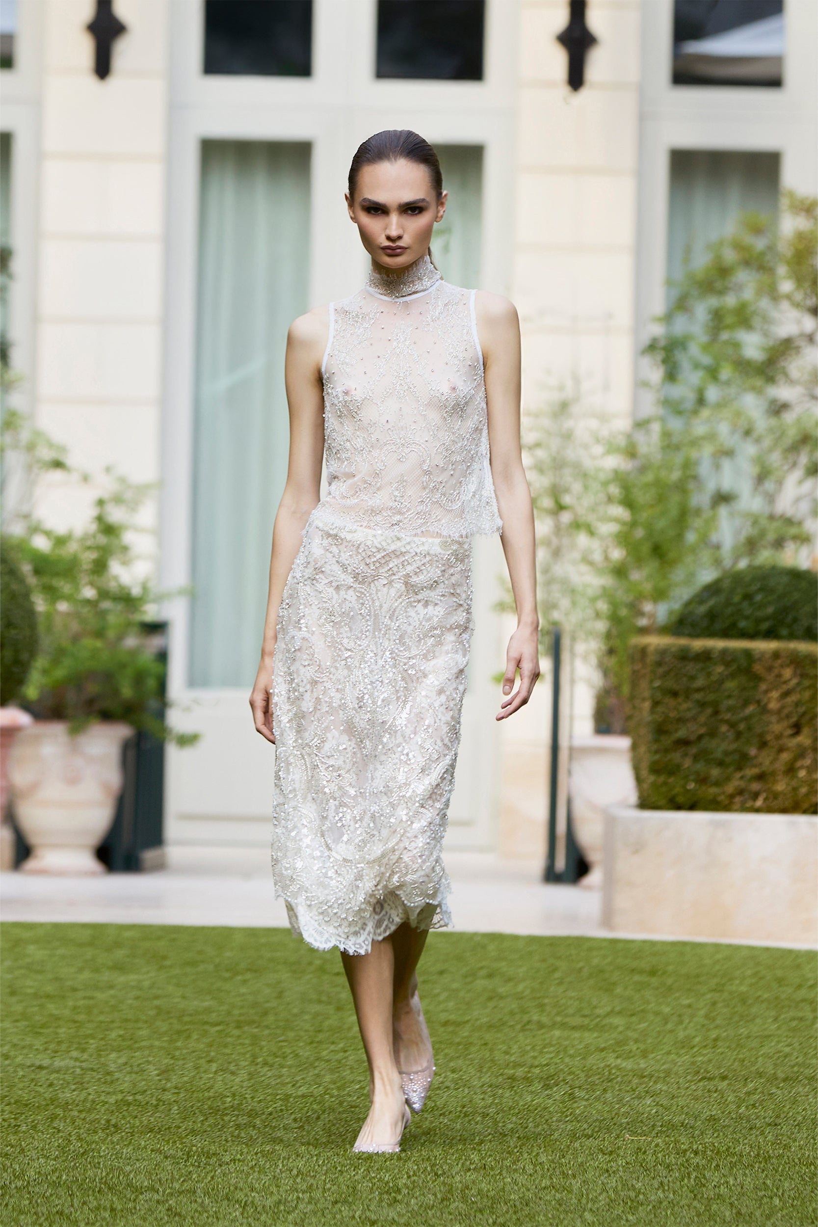 A model in a sleeveless, beaded white dress walks on grass in an outdoor garden setting, surrounded by large potted plants and a light-colored building in the background.
