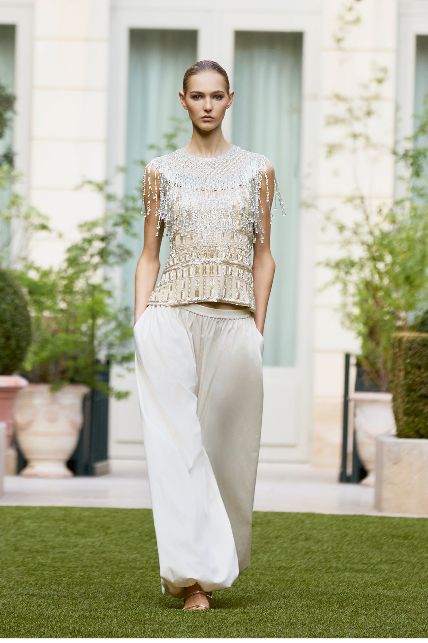 A model walks outdoors on grass in a sleeveless, beaded top with fringe and wide, white trousers. Blurred potted plants and tall windows add a natural touch to the background.
