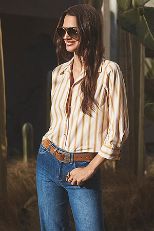 A woman wearing sunglasses, a striped button-up shirt, blue jeans, and a brown belt stands outdoors among plants, smiling with her hands in her pockets.