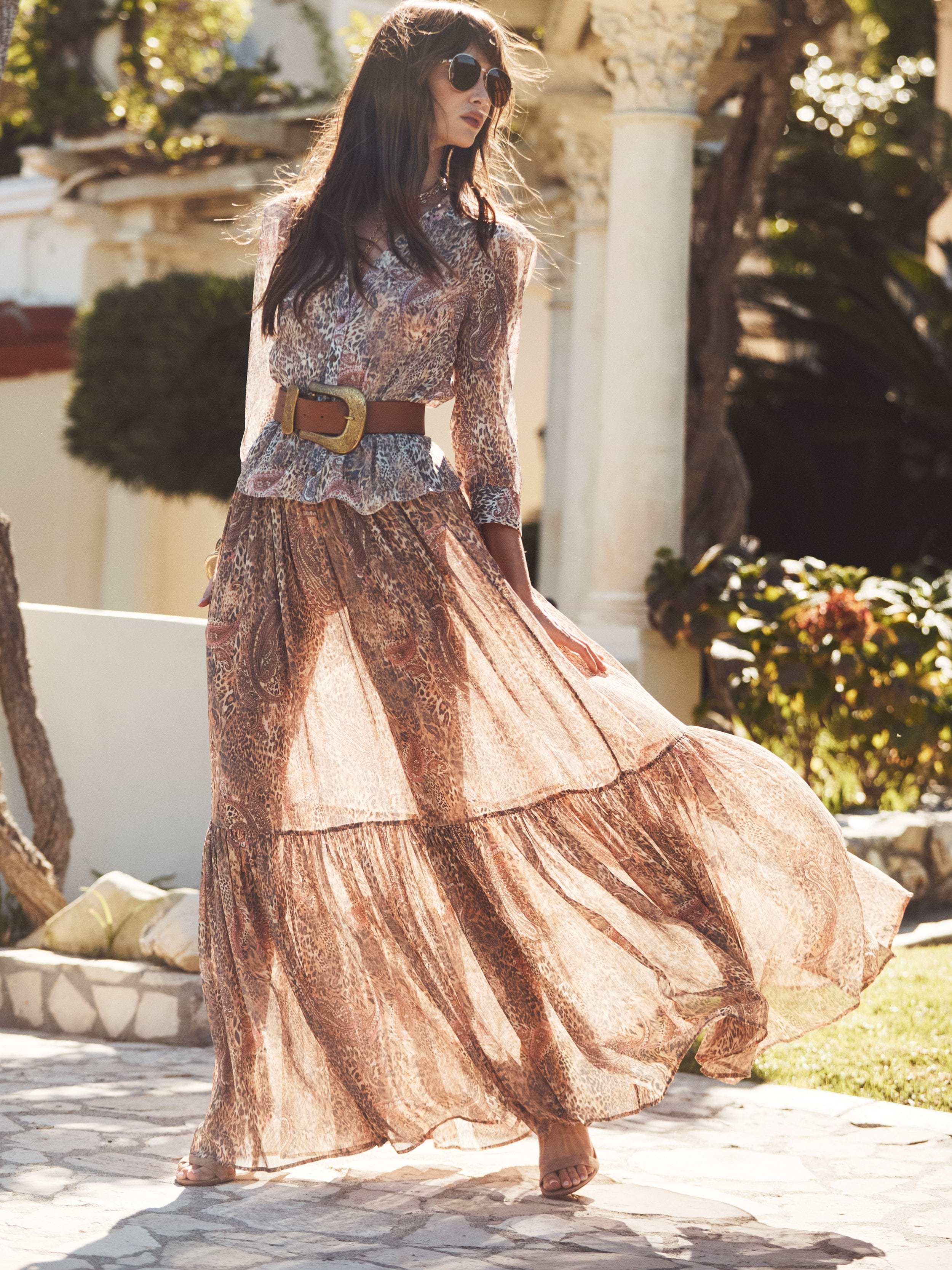 A woman in sunglasses wears a flowing, paisley-patterned long dress with a wide belt, standing outdoors on a sunny day with trees and a white building in the background.