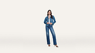 A woman stands against a plain white background, wearing a stylish blue denim jacket and matching jeans with black sandals. She faces forward with one hand in her pocket and the other adjusting her denim jacket lapel.
