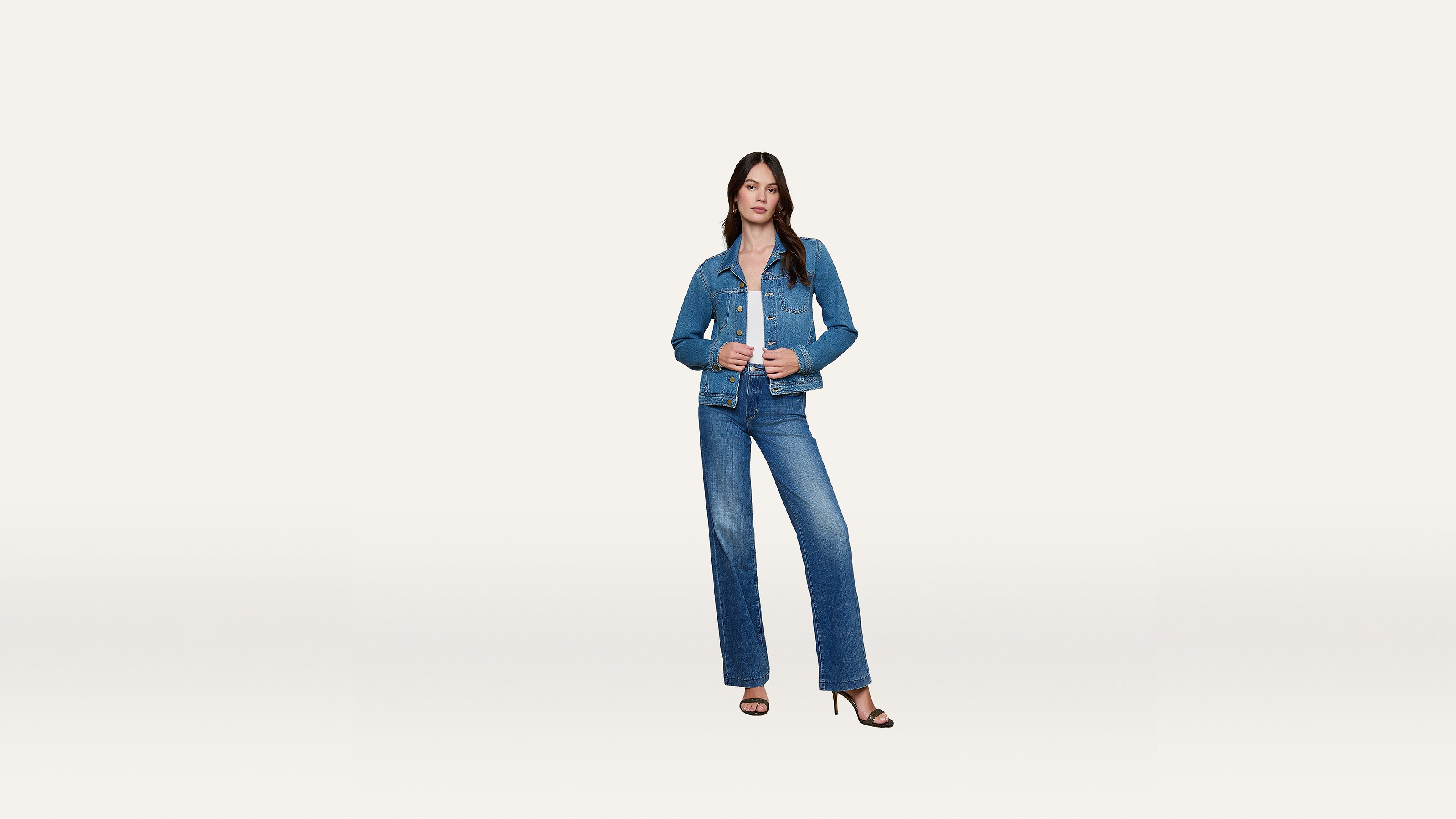 A woman stands against a plain white background, wearing a stylish blue denim jacket and matching jeans with black sandals. She faces forward with one hand in her pocket and the other adjusting her denim jacket lapel.