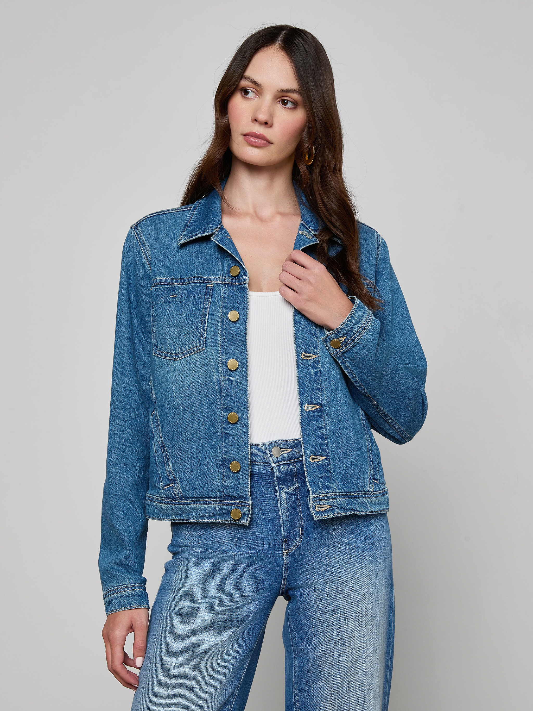 A woman wearing the featherweight denim Celine Jean Jacket from L’AGENCE over a white top and slim fit blue jeans stands against a plain, light background, looking slightly to her left with one hand holding the jacket collar.