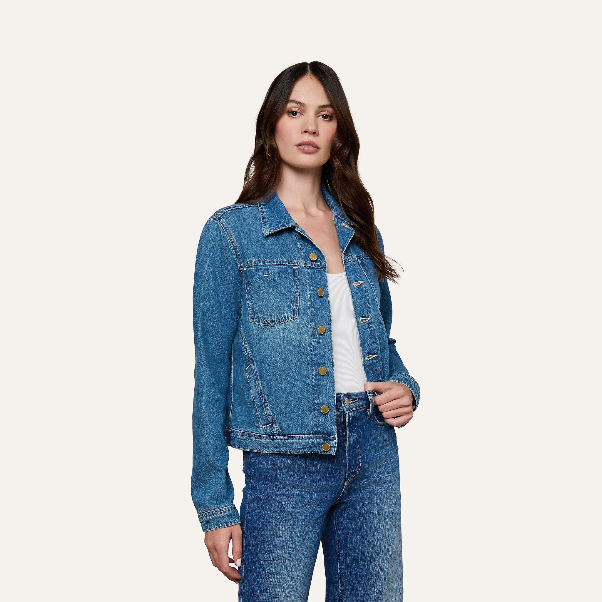 A woman wearing a slim fit Celine Jean Jacket over a white top and blue jeans stands against a plain, light background, looking towards the camera with her hand near the jacket’s buttons.