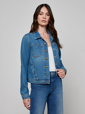 A woman wearing the Celine Jean Jacket in featherweight denim over a white top and slim fit blue jeans stands against a light grey background. She has long, loosely styled brown hair and a neutral expression.