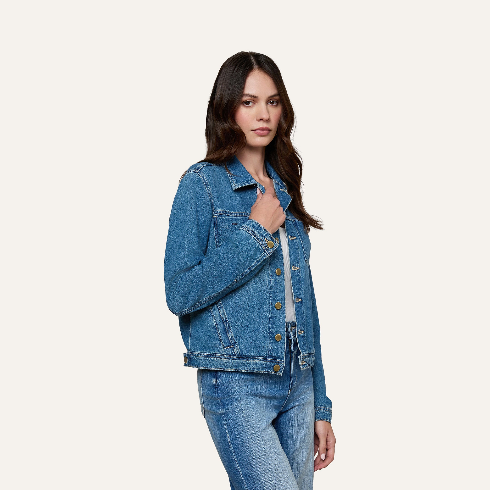 A woman wearing a featherweight denim Celine Jean Jacket over a blue top and slim fit jeans stands against a plain light background, looking at the camera with a neutral expression.