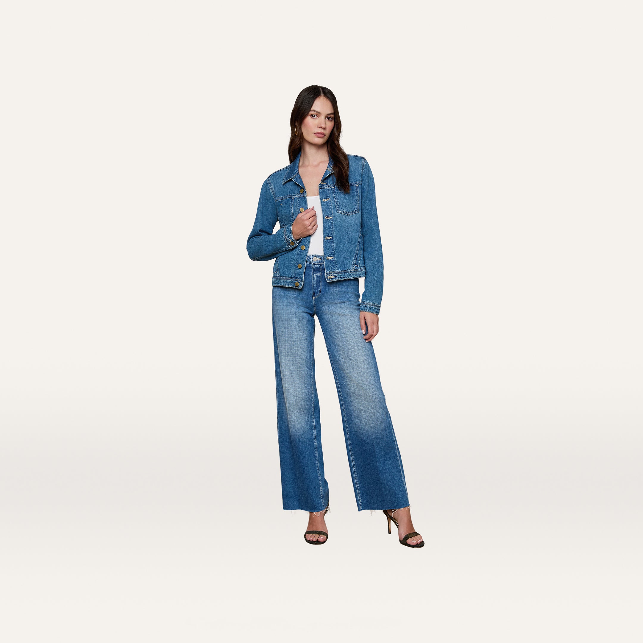 A woman stands against a plain white background, wearing the Celine Jean Jacket by L’AGENCE crafted from featherweight denim, paired with matching wide-leg jeans and black high-heeled sandals.