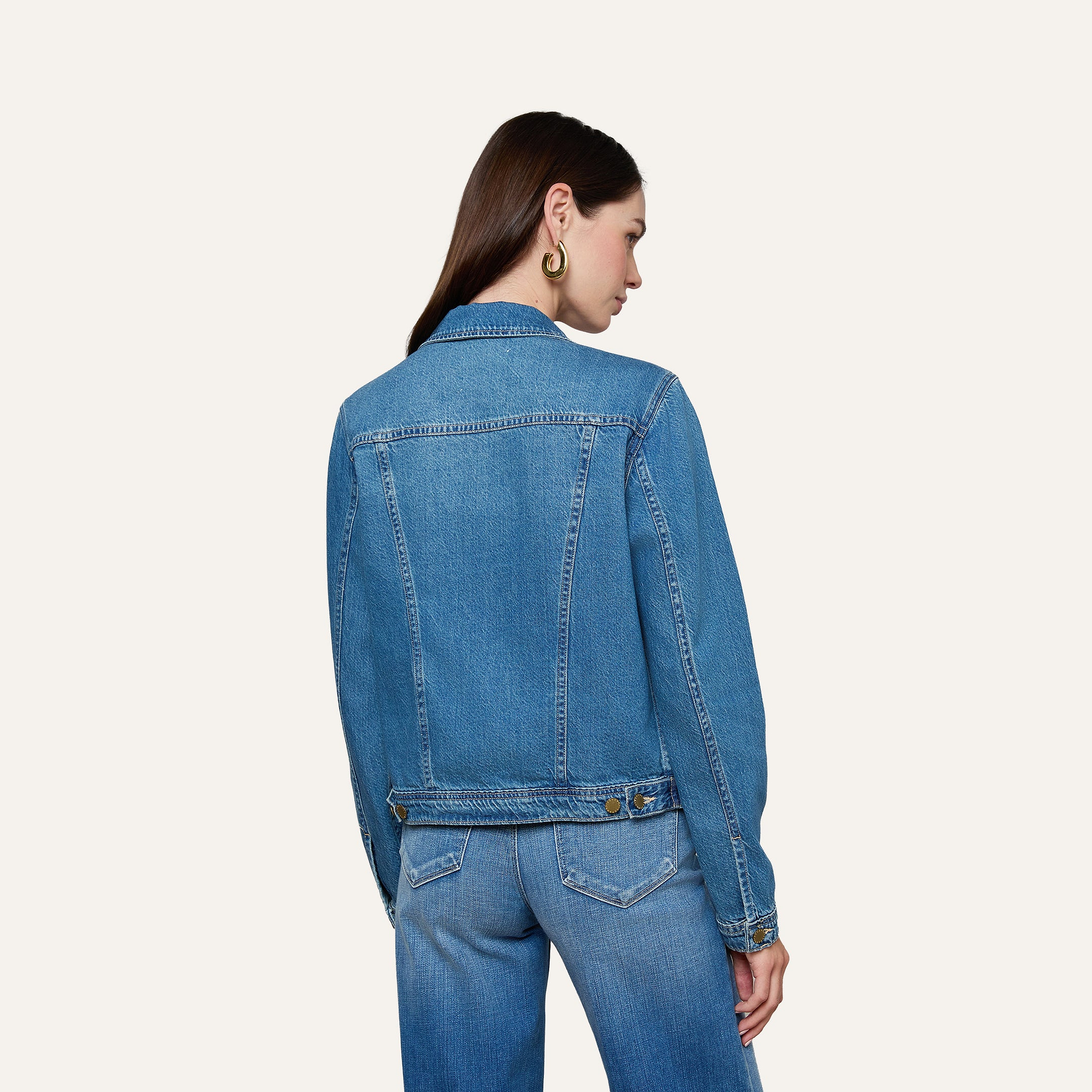 A woman with long brown hair, facing away from the camera against a plain light background, wears a featherweight denim Celine Jean Jacket and slim fit jeans.