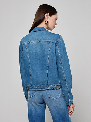 A woman with long brown hair, gold hoop earrings, and blue jeans stands facing away from the camera in a slim fit featherweight Celine Jean Jacket against a plain light gray background.