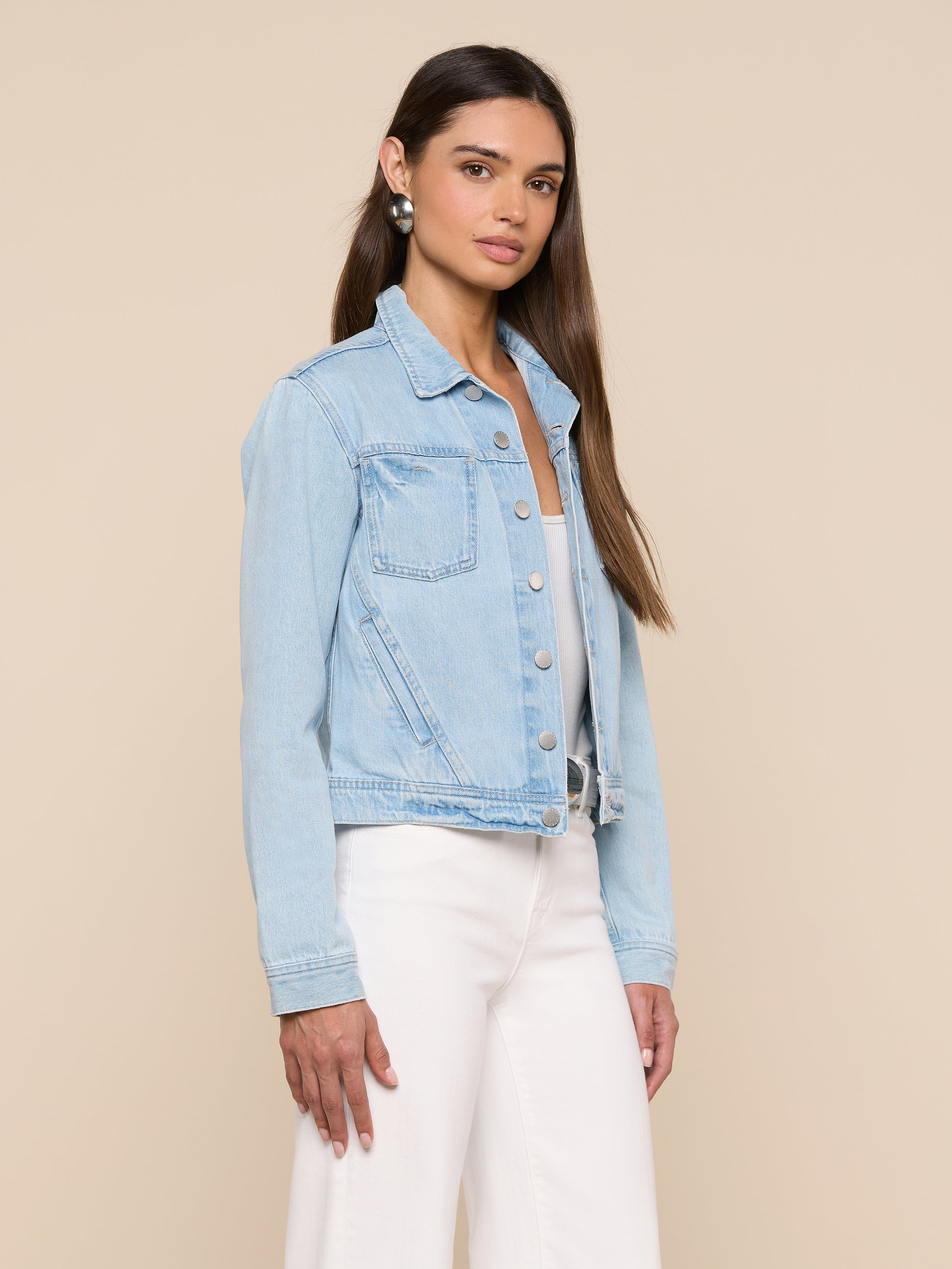 A woman with long brown hair wears a slim fit Celine Jean Jacket in light wash denim over a light top and white pants, standing against a plain beige background.