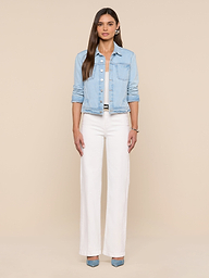 A woman with long brown hair and a neutral expression stands against a beige background, wearing the Celine Jean Jacket in light wash denim, white high-waisted wide-leg pants, and blue heels. Her arms are relaxed by her sides.