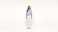 Woman stands facing forward against a plain white background, wearing a light denim jacket, white top, white wide-leg pants, and blue shoes. Her arms are relaxed by her sides and she has long, straight brown hair.