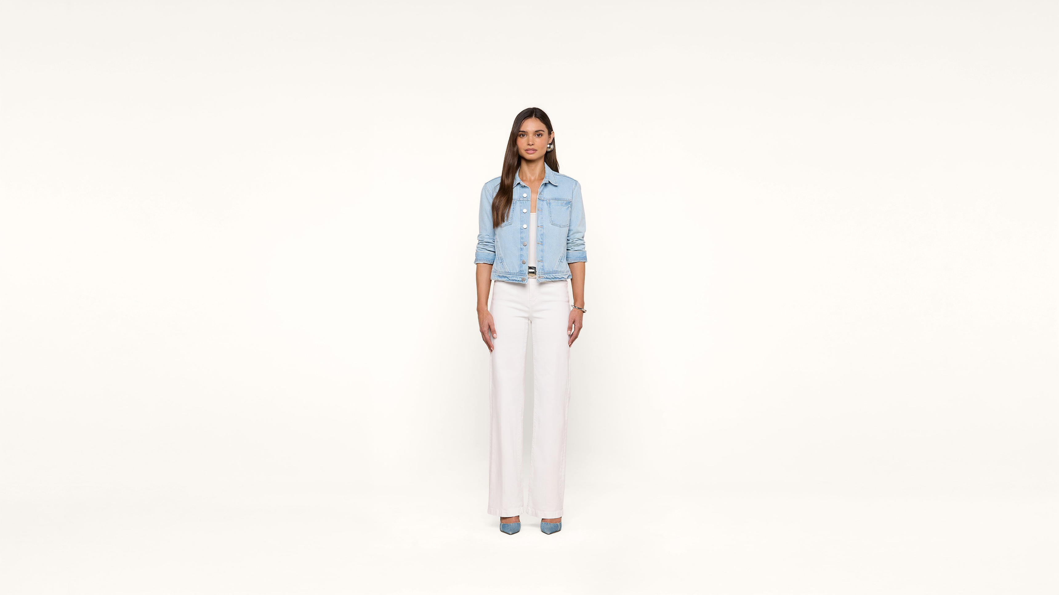 Woman stands facing forward against a plain white background, wearing a light denim jacket, white top, white wide-leg pants, and blue shoes. Her arms are relaxed by her sides and she has long, straight brown hair.