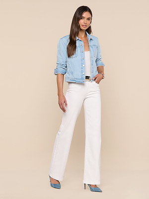 Woman with long hair stands confidently against a beige background, wearing the Celine Jean Jacket in light wash denim over a white top, paired with white high-waisted wide-leg pants, blue heels, and a black belt.