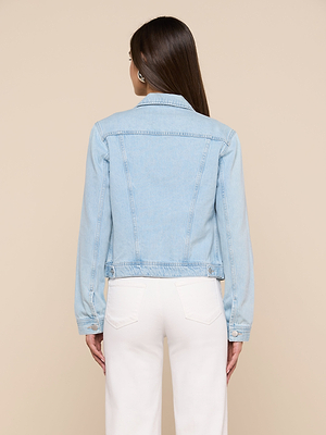 A woman with straight brown hair faces away, wearing a slim fit Celine Jean Jacket in light wash denim and white pants, set against a plain beige background.