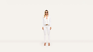 A woman wearing sunglasses, a white jacket, white top, white pants, and tan heels poses stylishly against a plain, light background.