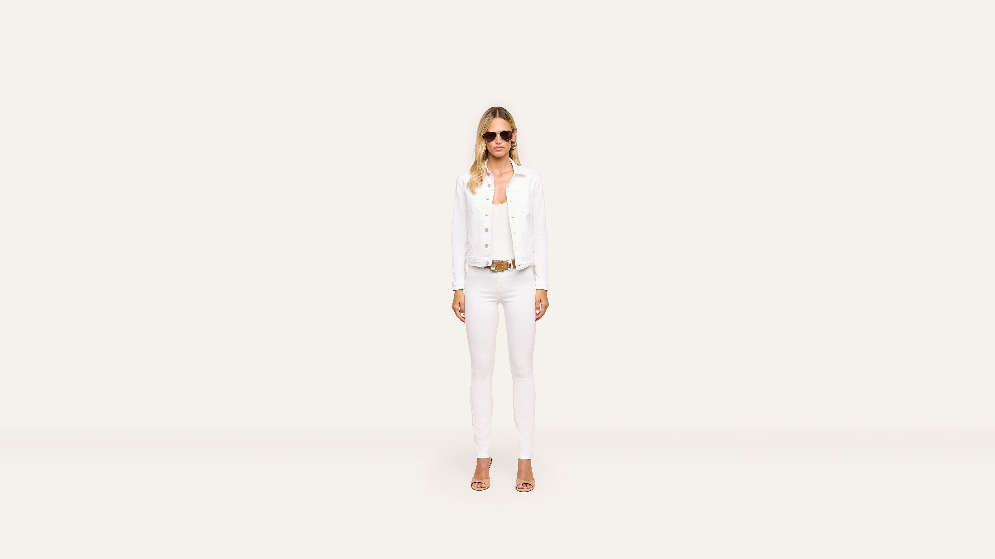 A woman wearing sunglasses, a white jacket, white top, white pants, and tan heels poses stylishly against a plain, light background.