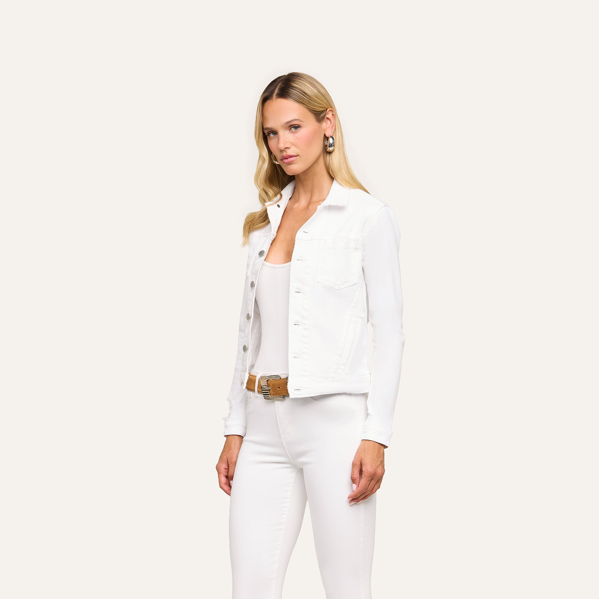 A woman with long blonde hair wears the Celine Jean Jacket in optic white and white pants with a brown belt, standing confidently against an off-white background.