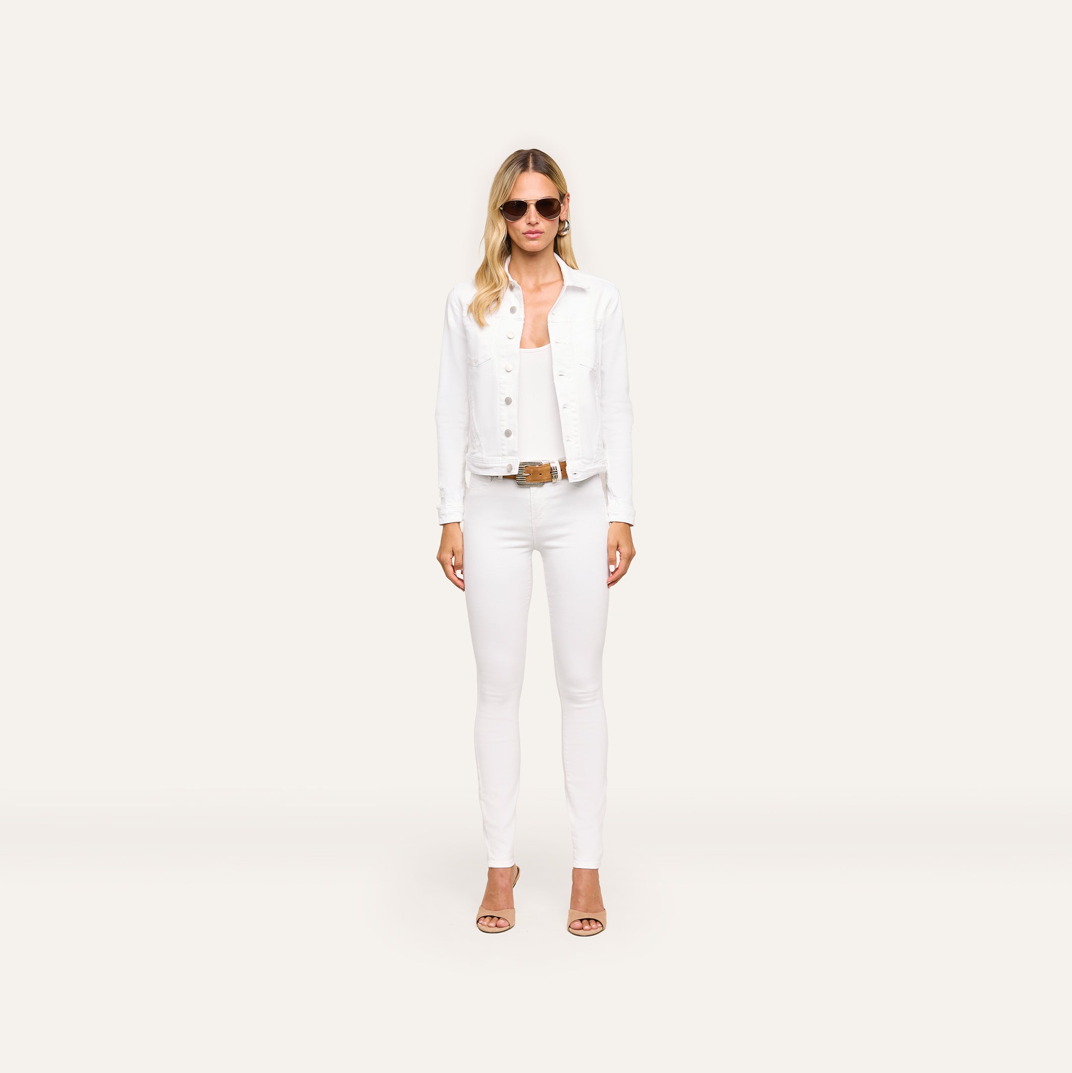 A woman with long blonde hair wears sunglasses, the Celine Jean Jacket in optic white with a relaxed fit, white pants, and tan heels, standing against a plain light background.