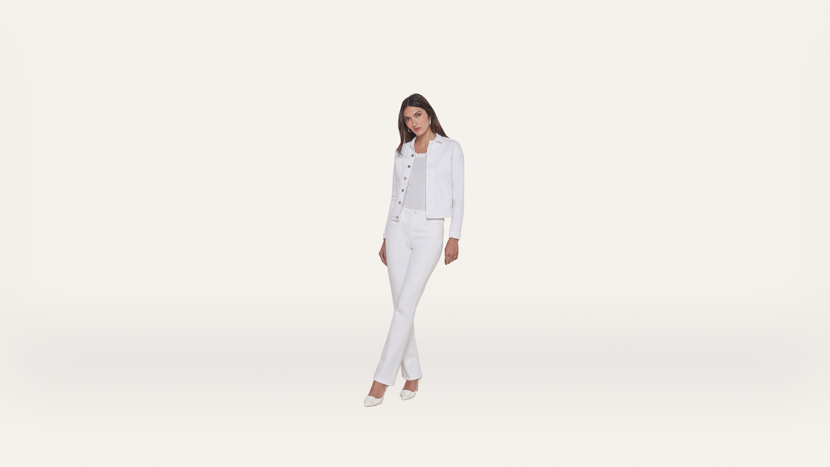 Woman standing and posing in all-white clothing: a denim jacket, top, jeans, and heels, against a plain white background.