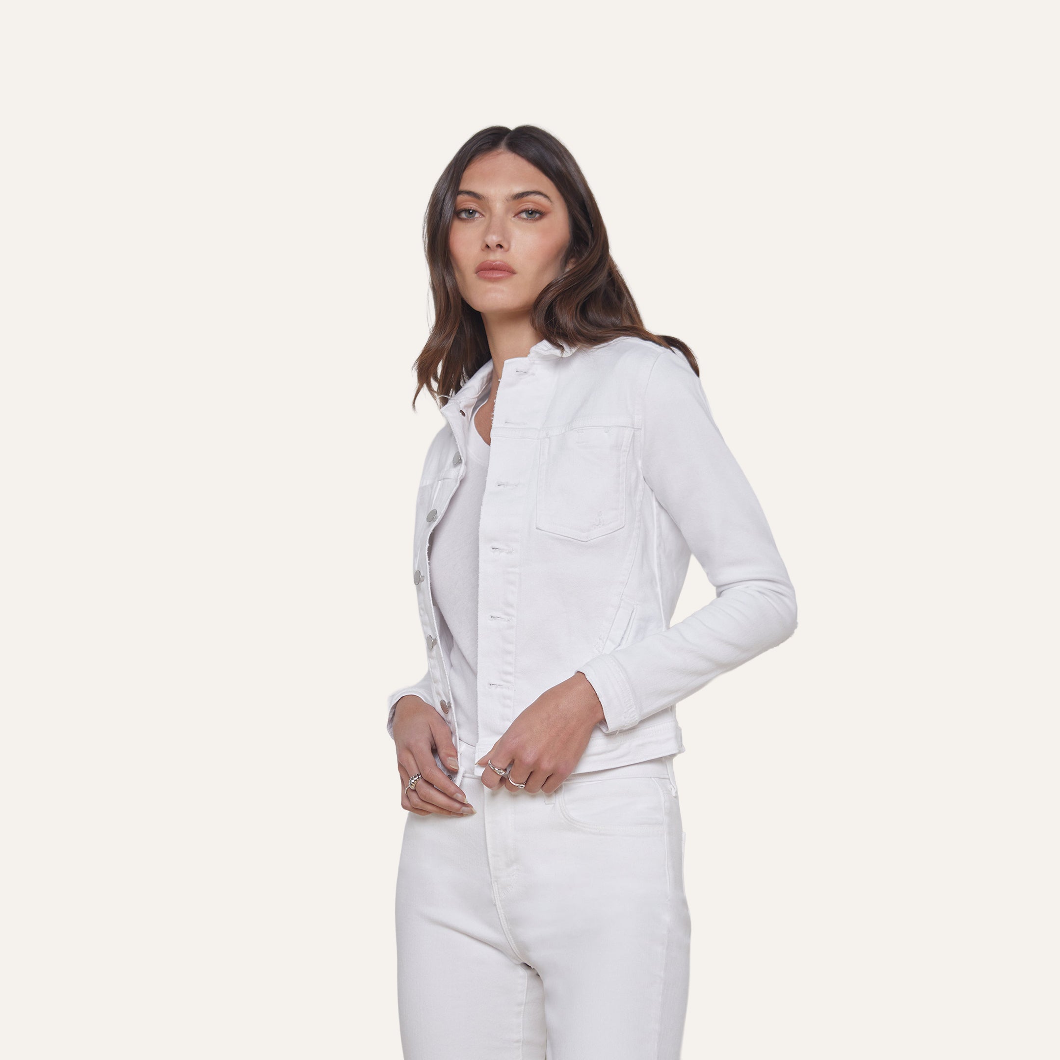 A woman with long brown hair wears the Celine Jean Jacket in optic white, featuring a relaxed fit, layered over a light top and matching white jeans, standing against a plain off-white background.