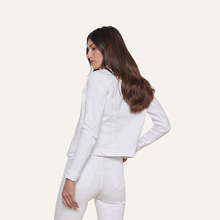 A woman with long brown hair is standing sideways and looking down. She is wearing a white denim jacket and white jeans against a plain, light background.