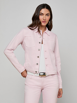 A woman with long brown hair poses against a plain gray background, wearing an all-cotton Celine Jean Jacket by L’AGENCE with matching pastel lilac pants, a white top, and belt, one hand in her pocket.