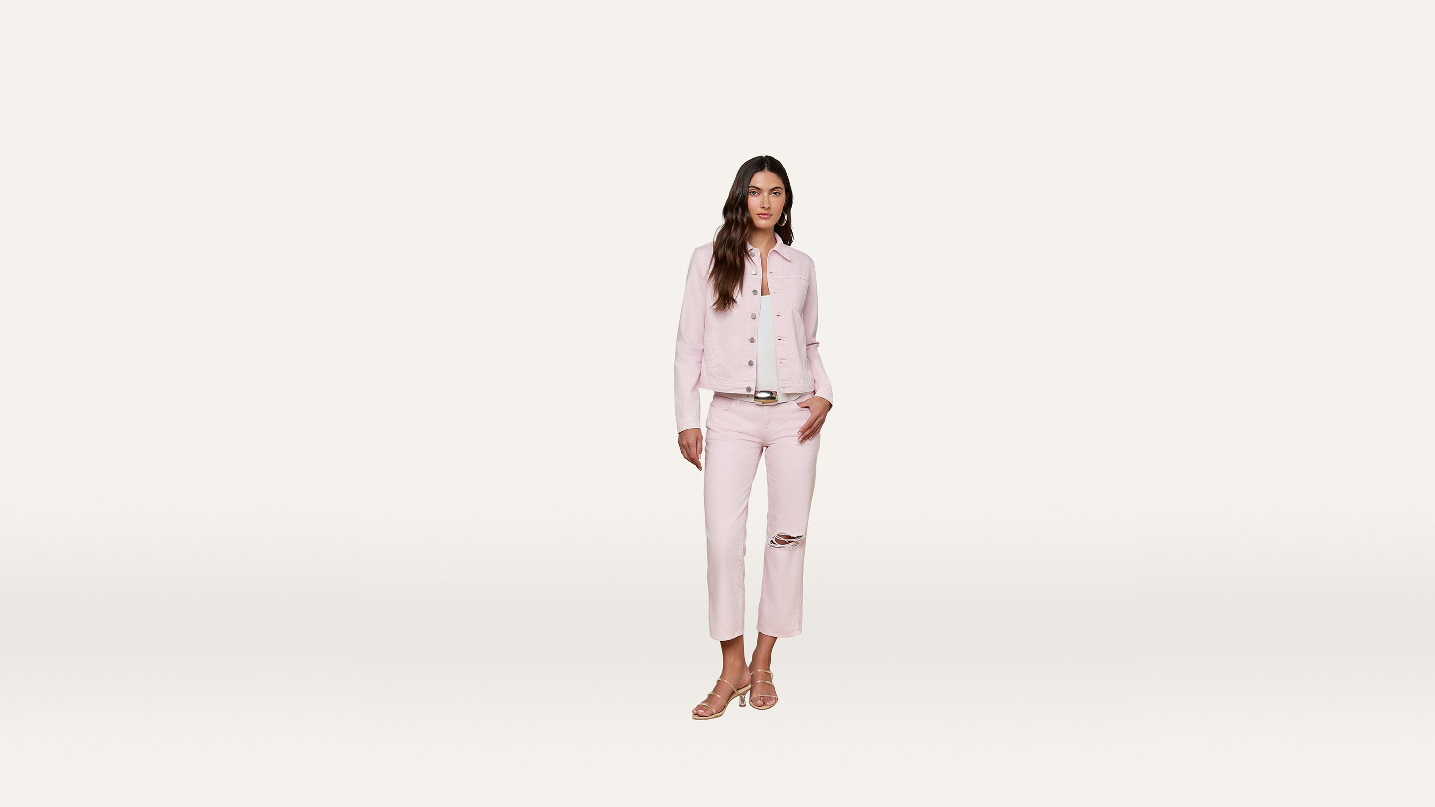Woman wearing a light pink jacket and matching cropped pants with a knee rip, white shirt, and sandals, standing against a simple white background.