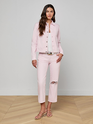 A woman with long, wavy dark hair and a neutral expression stands indoors against a light gray wall wearing a Celine Jean Jacket and matching lilac denim pants with a knee rip, white top, metallic belt, and sandals.