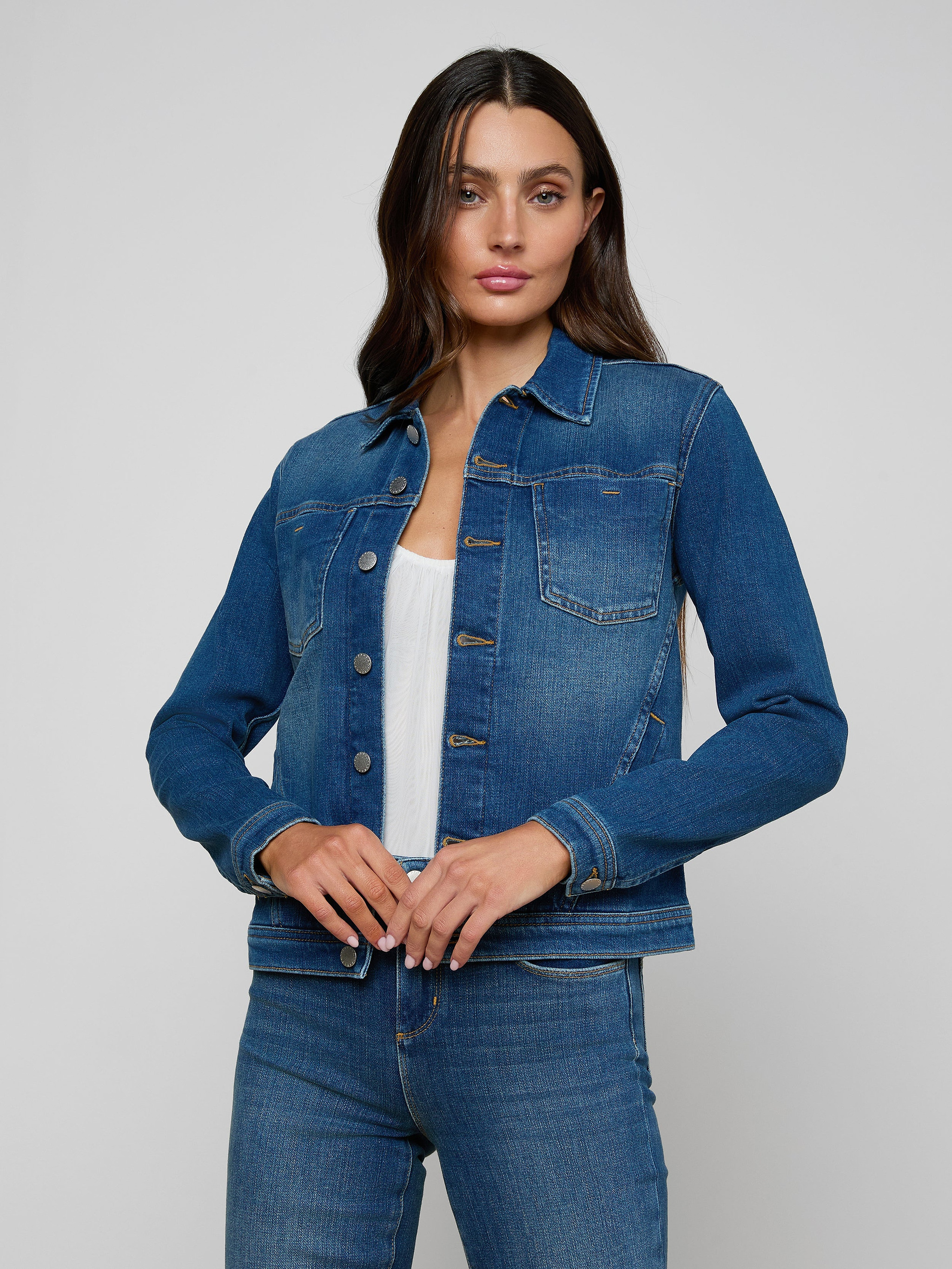 Woman wearing a slim fit Celine Jean Jacket by L’AGENCE over a white top and matching blue jeans, standing against a plain light background, looking forward with one hand adjusting the jacket.