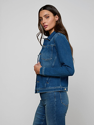 A woman with long brown hair wears a Celine Jean Jacket and slim fit jeans, standing sideways against a plain light background.