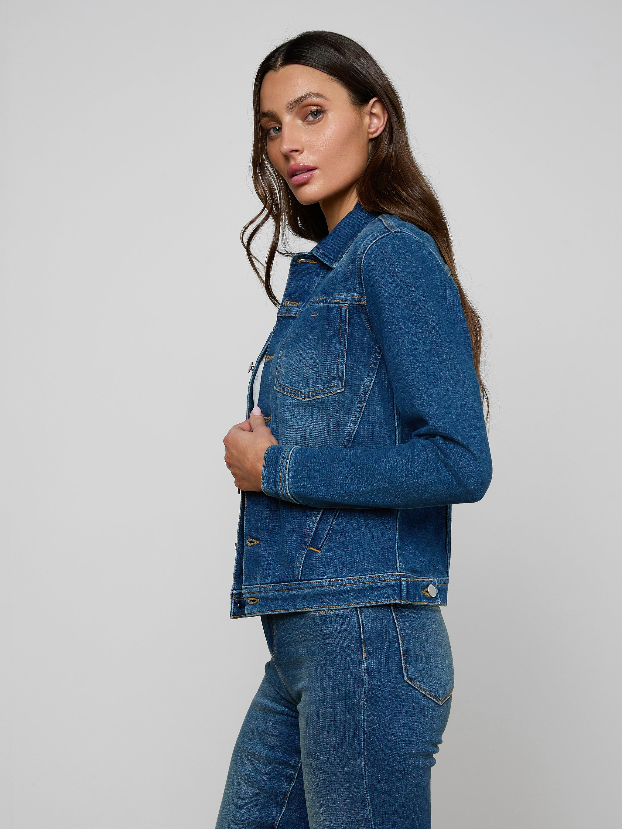 A woman with long brown hair wears a Celine Jean Jacket and slim fit jeans, standing sideways against a plain light background.