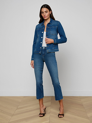 A woman stands indoors on a wooden floor wearing the slim fit Celine Jean Jacket and matching blue jeans with a white top and black heeled sandals, posing against a plain light-colored wall.