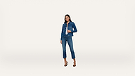 A woman stands against a plain white background wearing a blue denim jacket and matching blue jeans with black high-heeled sandals.
