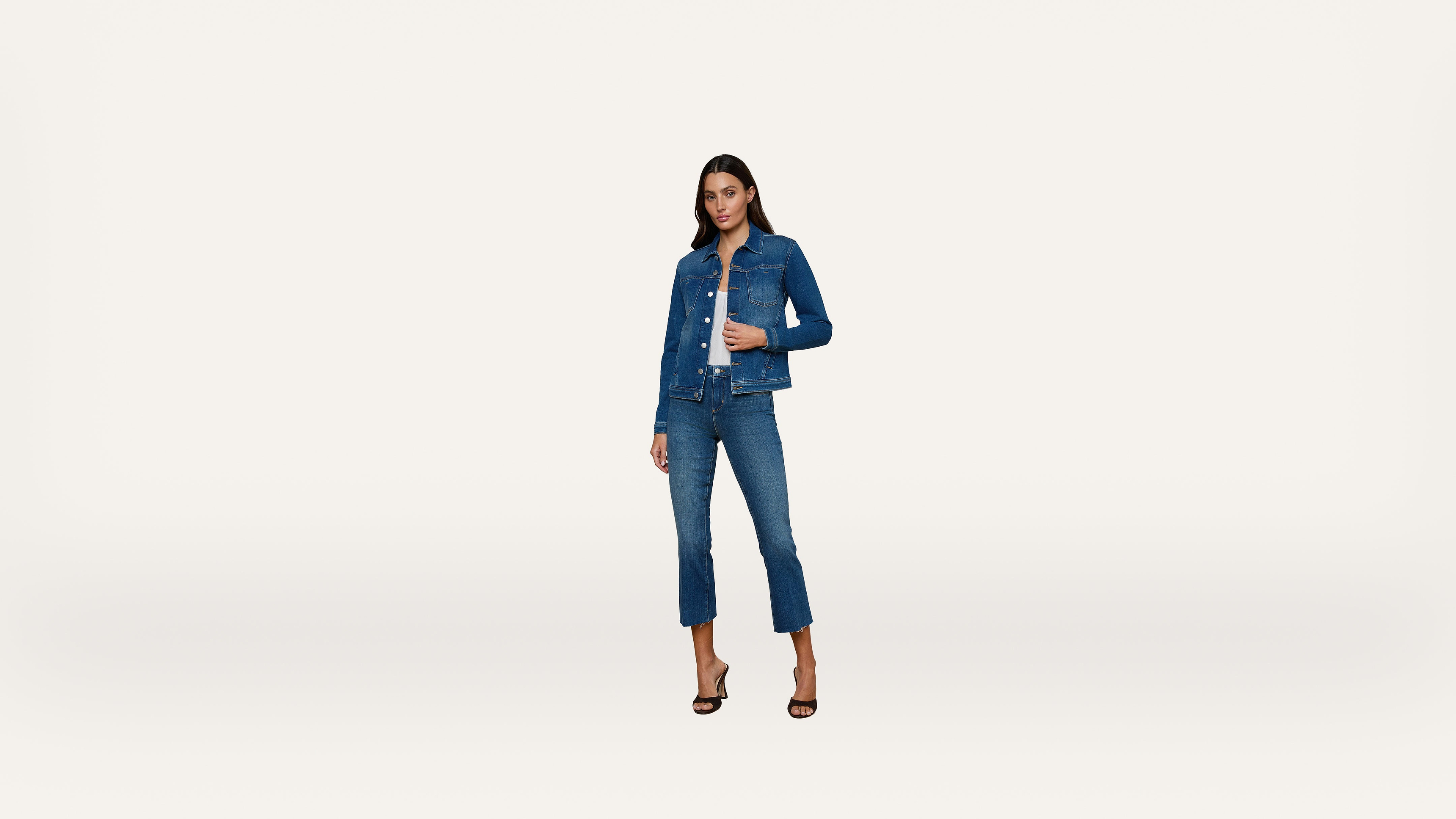 A woman stands against a plain white background wearing a blue denim jacket and matching blue jeans with black high-heeled sandals.
