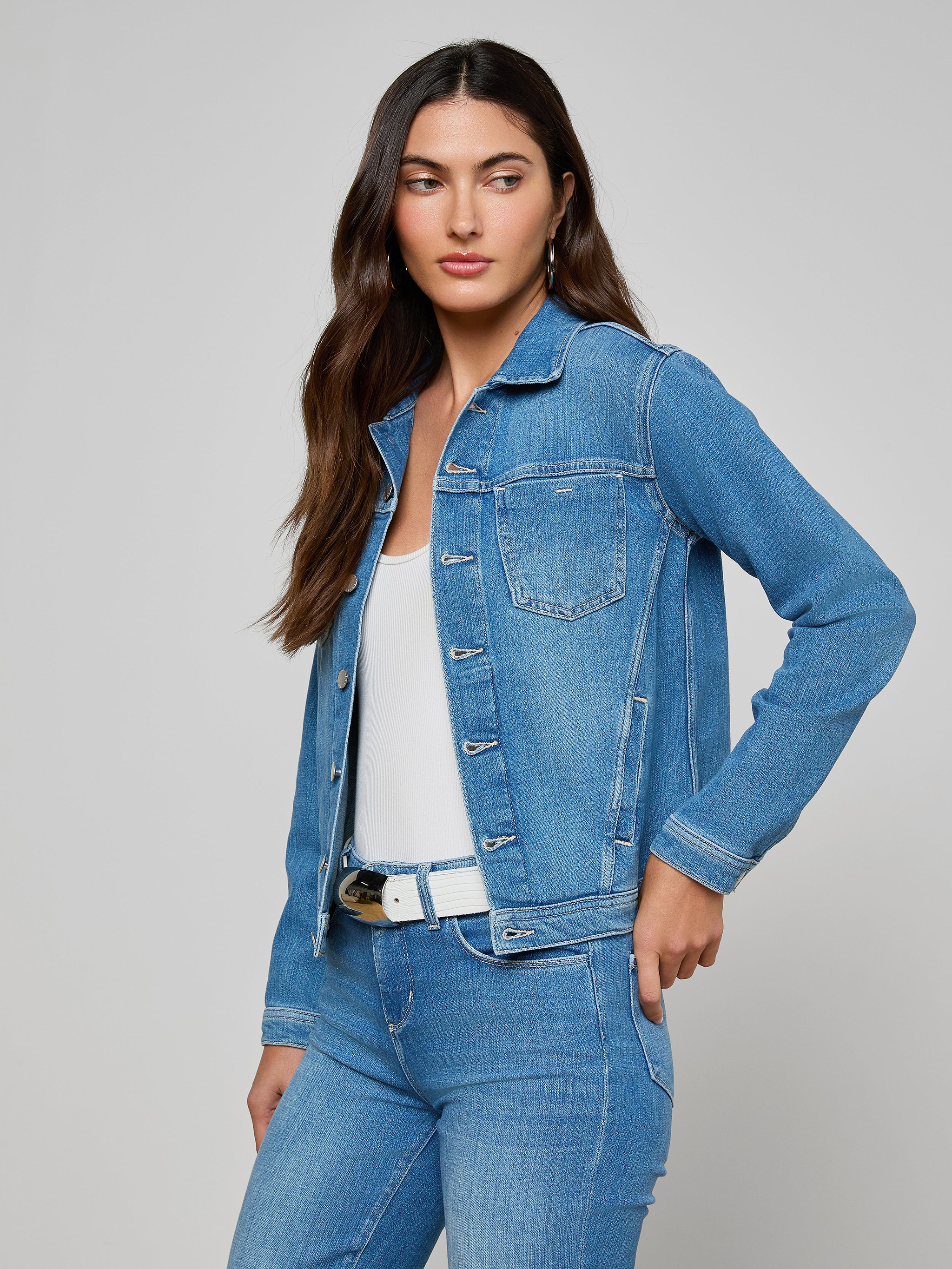 A woman wearing the Celine Jean Jacket in a slim fit with matching jeans over a white top stands with one hand in her pocket, looking to her left.
