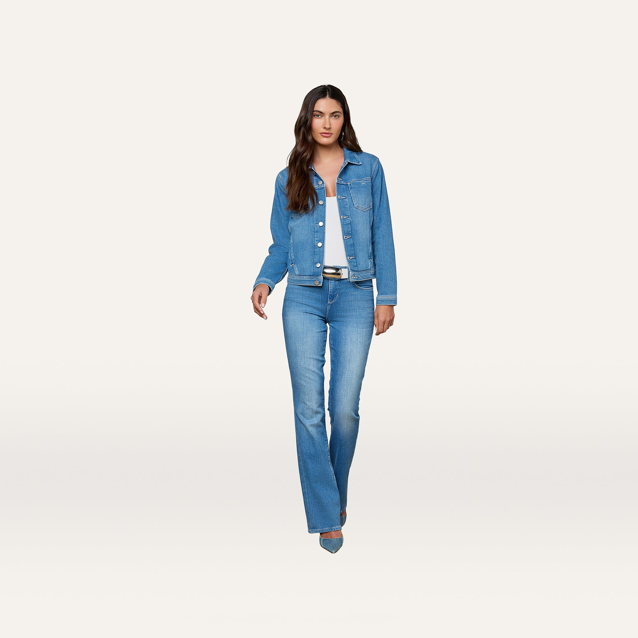 A woman stands against a plain white background wearing the Celine Jean Jacket by L’AGENCE and matching jeans over a white top, paired with blue heels. This versatile essential features a slim fit that perfectly complements her modern, polished look.