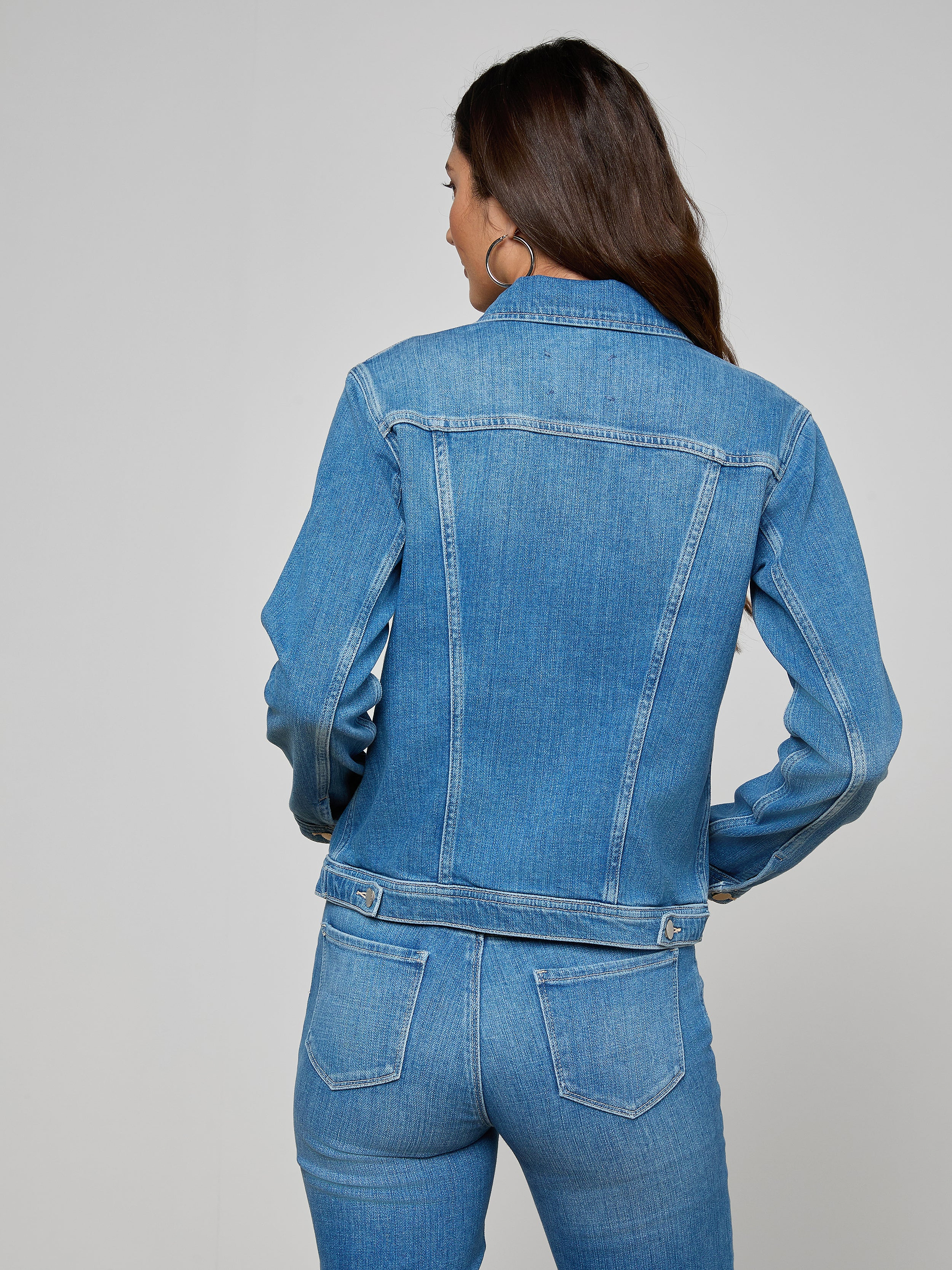 Wearing the Celine Jean Jacket, a woman with long brown hair, seen from behind, pairs this slim fit denim piece with matching blue jeans against a plain, light-colored background—showcasing the versatility of this essential.