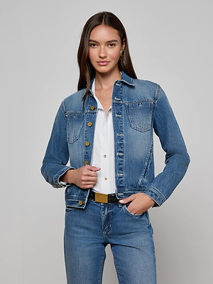 A woman with long brown hair wears the L'AGENCE Celine Jean Jacket over a white shirt and blue jeans, standing with one hand on her hip against a plain light gray background.