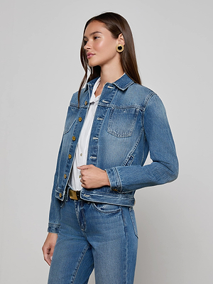 A woman with long brown hair wears the L’AGENCE Celine Jean Jacket in classic denim blue over a white shirt and slim fit jeans. She poses sideways, hand on the jacket's waist, accessorized with gold hoop earrings against a neutral background.
