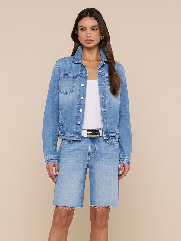 A woman stands against a plain light background wearing the Celine Jean Jacket from L’AGENCE in a light denim wash, styled over a white top and matching knee-length denim shorts.