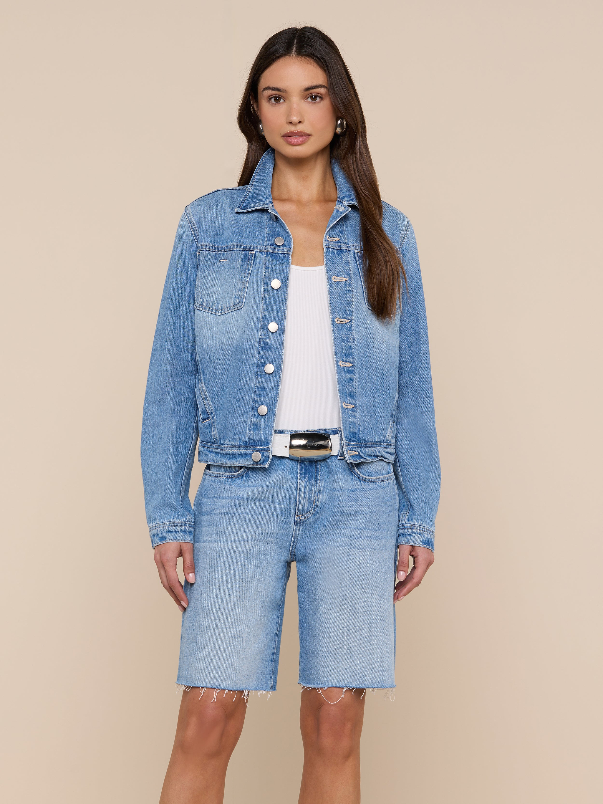A woman stands against a plain light background wearing the Celine Jean Jacket from L’AGENCE in a light denim wash, styled over a white top and matching knee-length denim shorts.