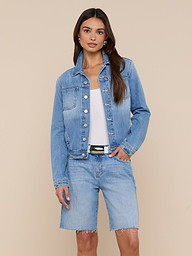 A woman wearing the Celine Jean Jacket in a light denim wash with matching knee-length denim shorts, a white top, and a black belt stands with one hand in her pocket against a plain background.