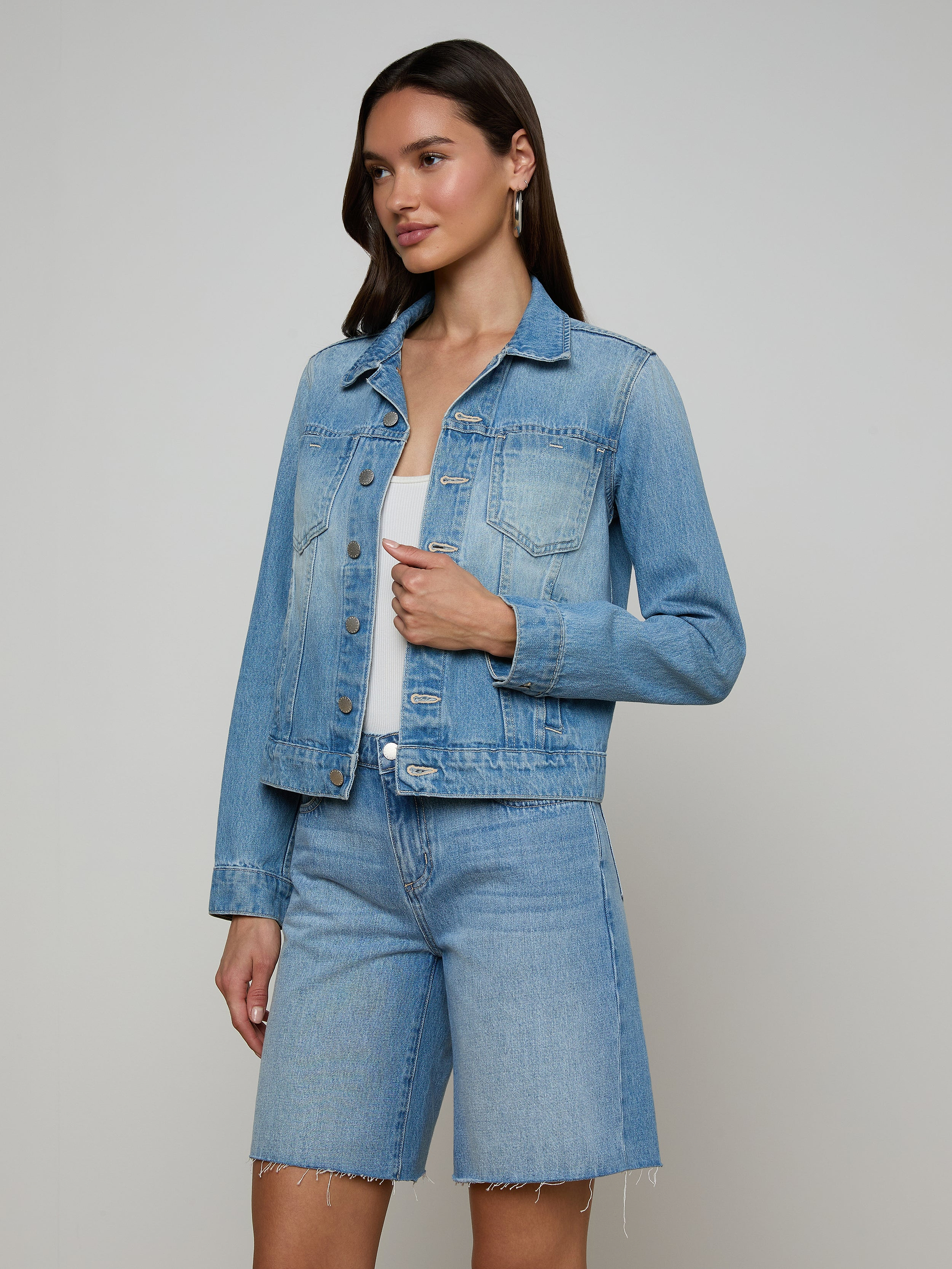 A woman stands in a light denim wash Celine Jean Jacket and matching slim fit, knee-length denim shorts against a plain, light background. She has long brown hair and is looking slightly to the side.