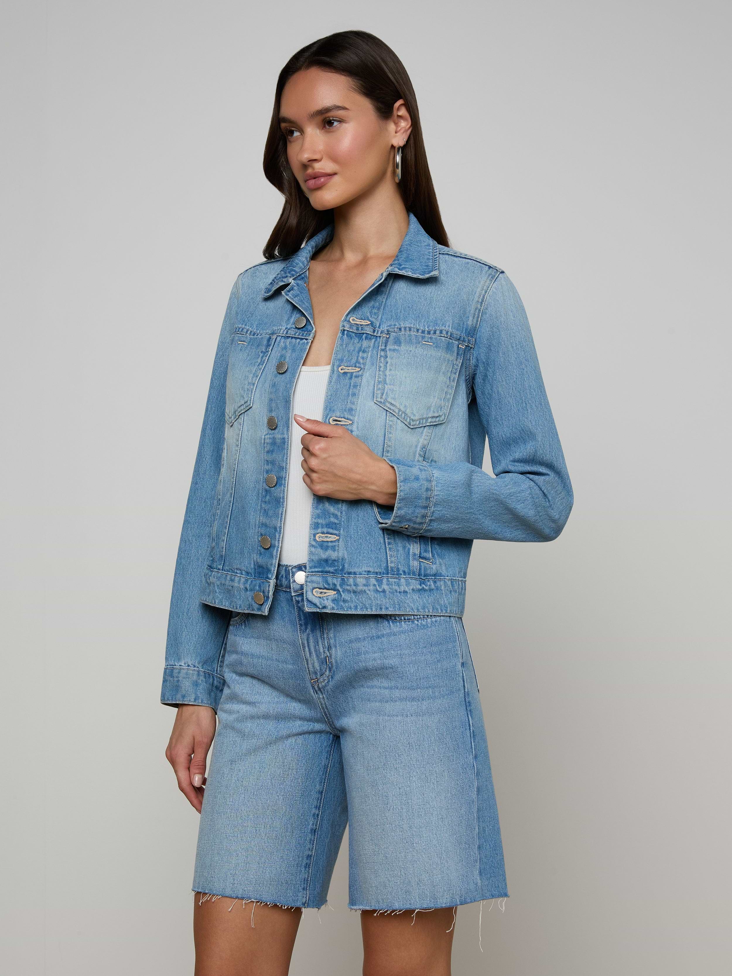 A woman stands in a light denim wash Celine Jean Jacket and matching slim fit, knee-length denim shorts against a plain, light background. She has long brown hair and is looking slightly to the side.
