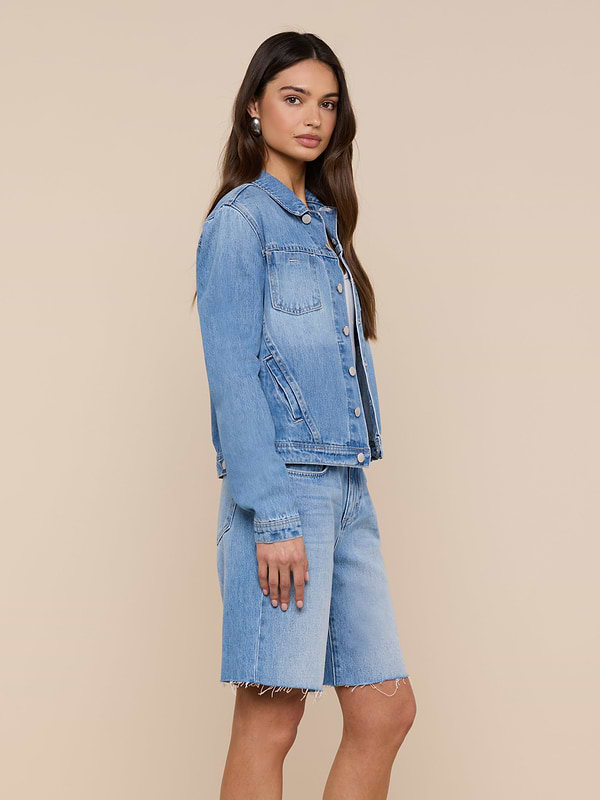 A woman stands sideways, wearing a slim fit light denim wash Celine Jean Jacket and matching knee-length denim shorts against a plain white background.