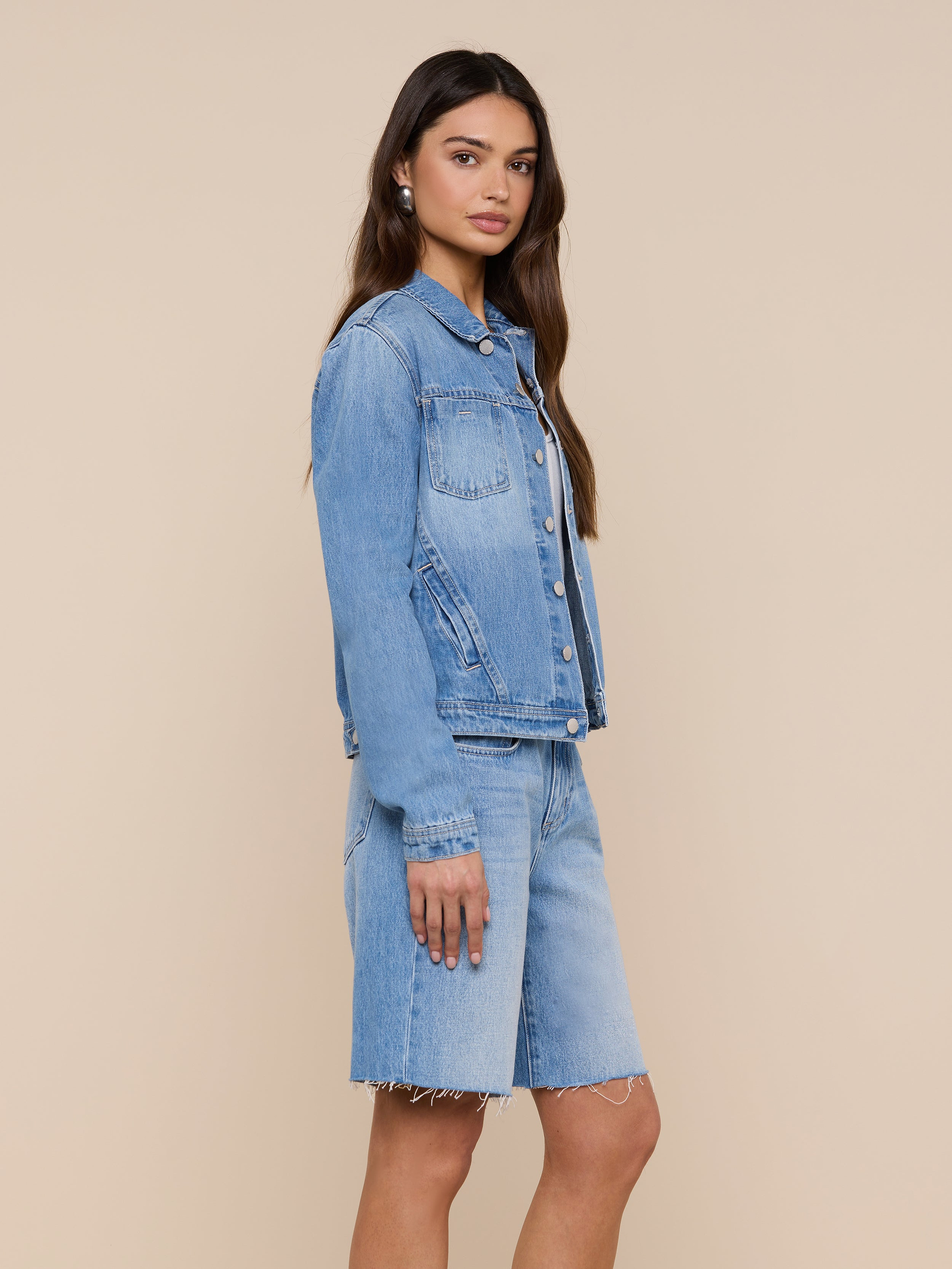 A woman stands sideways, wearing a slim fit light denim wash Celine Jean Jacket and matching knee-length denim shorts against a plain white background.