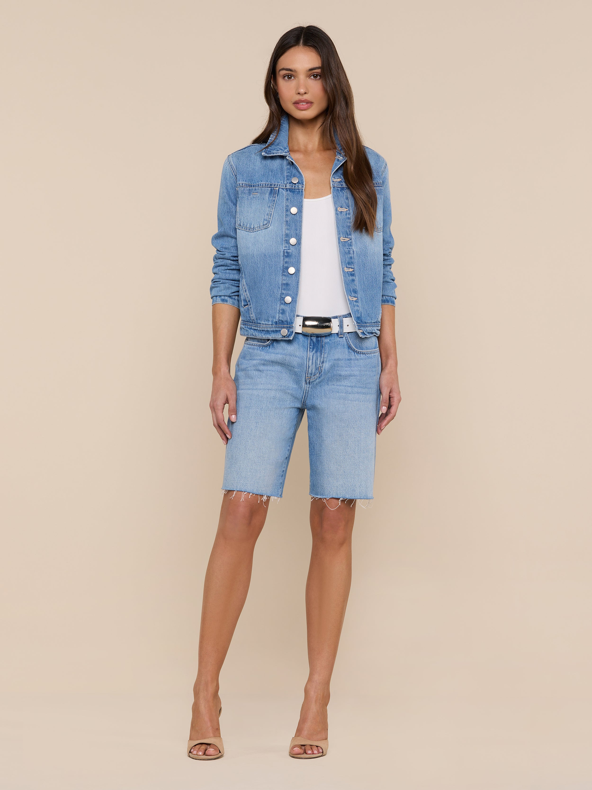 A woman stands facing forward, wearing the Celine Jean Jacket by L’AGENCE in a light denim wash, a white top, slim fit light blue knee-length denim shorts, a black belt, and beige heels against a plain white background.