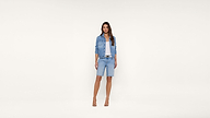 Woman standing against a plain white background, wearing a blue denim jacket, light blue top, denim shorts, and nude heels. She has long, straight hair and a neutral expression.